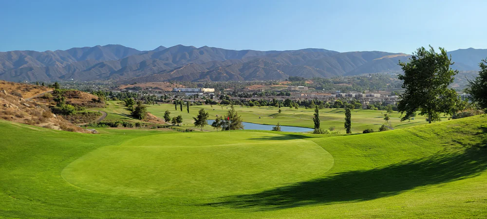 Expansive golf course landscape with a vibrant green putting green in the foreground, a lake in the middle ground, and a range of mountains stretching across the horizon.